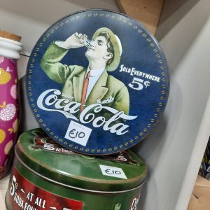 a vintage round coke tin with an image of a man in a green fedora and green jacket drinking coke. the tin is navy and the coca cola wording is white. there is also a price of 5 cent on it in keeping with the vintage theme