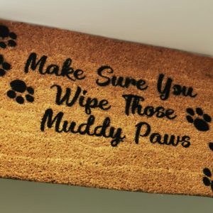 a brown doormat with black writing which says make sure you wipe those muddy paws. there are 3 black pawprints on one side of the writing and 3 on the other